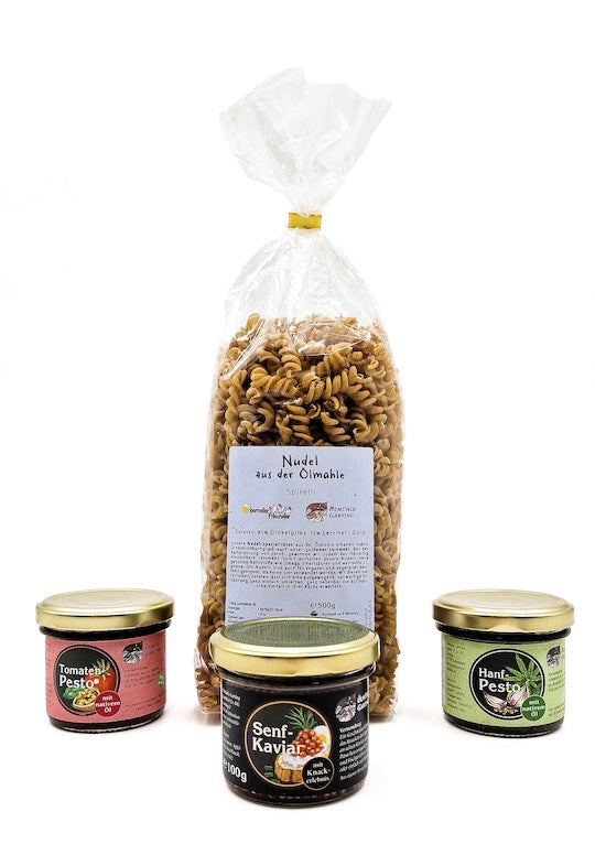 Eine Packung Spiralnudeln aus der Ölmühle Garting, umgeben von drei Feinkost-Gläsern mit Tomatenpesto, Hanfpesto und Senf-Kaviar. Die Produkte stehen auf weißem Hintergrund und zeigen das Sortiment an regionaler Pasta und hausgemachten Delikatessen.