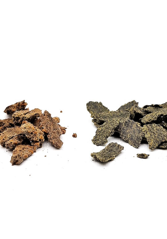 Zwei Haufen unterschiedlicher Bio-Presskuchen der Ölmühle Garting auf weißem Hintergrund – links ein hellbrauner, rechts ein dunkelgrüner Presskuchen aus kaltgepressten Ölsaaten, verwendet als hochwertiges Futtermittel.