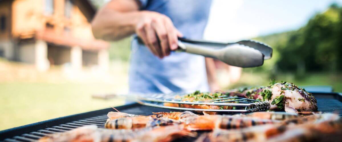 Person am Grill mit Grillzange, auf dem Grill ist Fleisch zu sehen mit Marinade