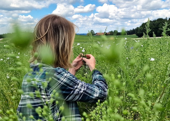 Frau mit blonden Haaren und einem grünkariertem Hemd auf einem Leindotterfeld, im Hintergrund Himmel mit weißen Wolken