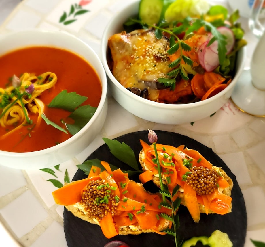 Links rote Suppe, Mitte Brot mit Karotte und Senfkaviar und hinten ein gemischter Salat mit Kräutern auf einer Tischdecke