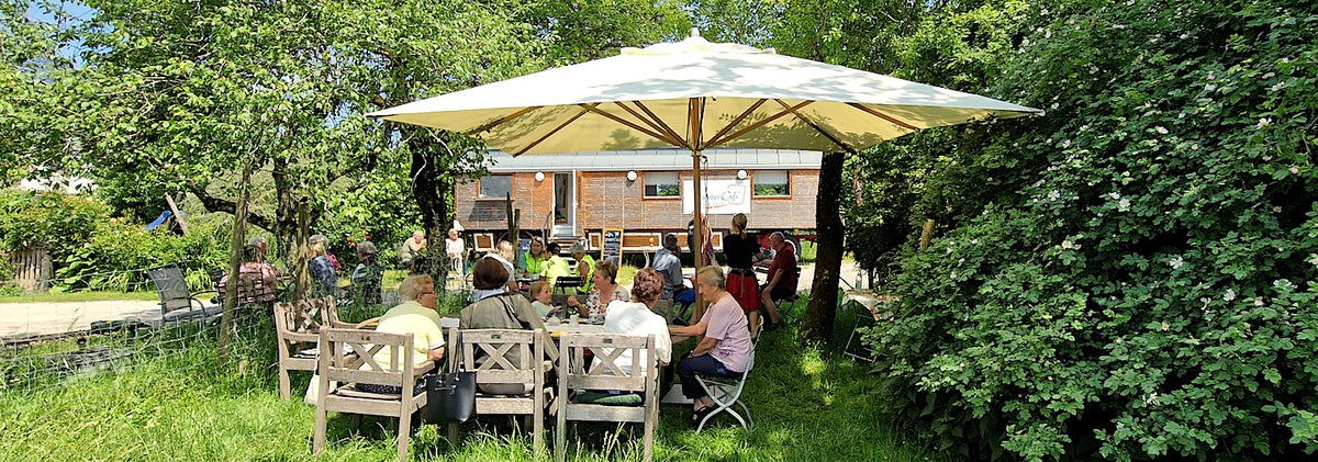 Das Wandercafe und der Garten mit Sonnenschirm und Gästen im Vordergrund, Wandercafe im Hintergrund